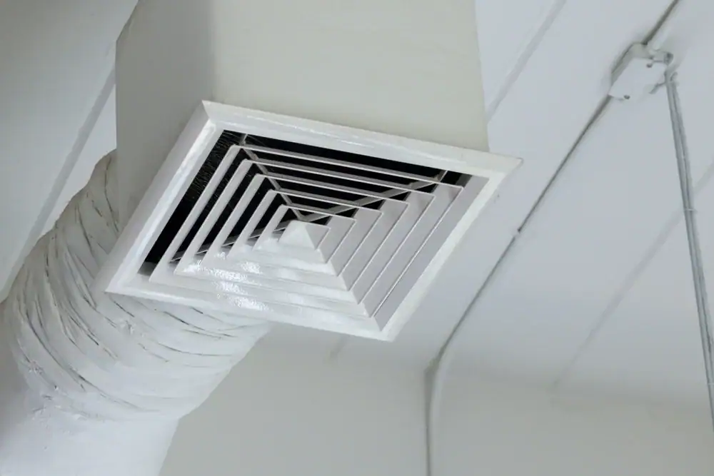 A close-up view of a white, square ceiling air vent attached to a duct, with geometric slats arranged in a concentric pattern—a clean finish often seen after professional carpet cleaning in Houston County, TX. Exposed pipes line the white ceiling background.