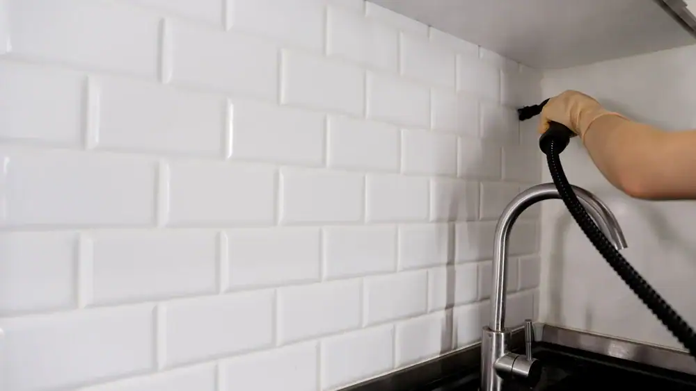 A person wearing a glove uses a steam cleaner to clean white subway tile backsplash behind a kitchen sink with a silver faucet, showcasing the same care and attention found in top-rated carpet cleaning Houston County, TX services.