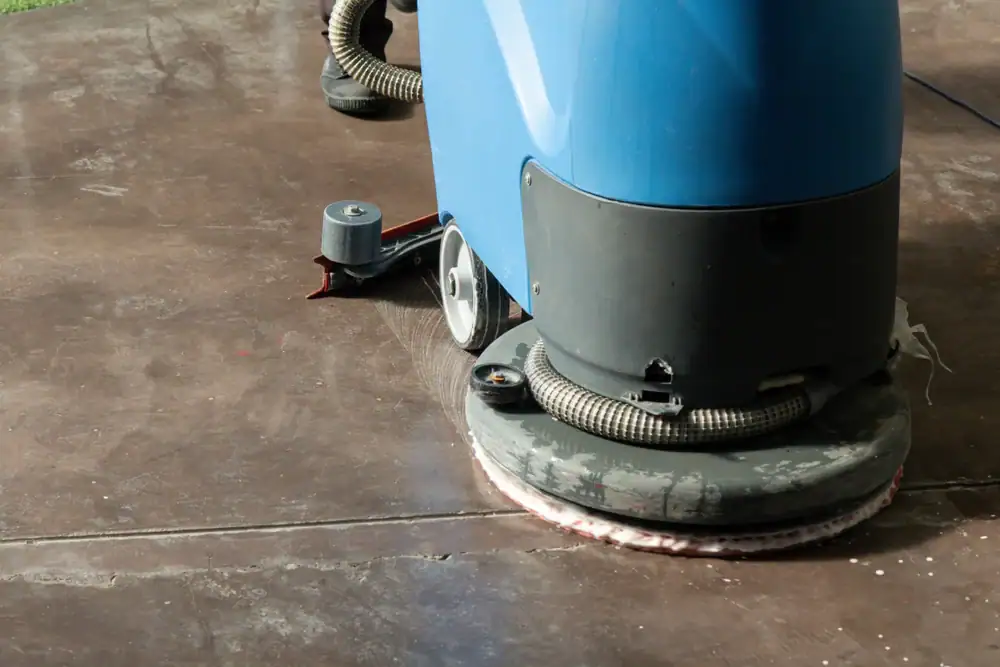 A close-up of a floor cleaning machine scrubbing a brown surface, with part of the operator’s shoes visible in the background, highlights professional carpet cleaning in Houston County, TX.