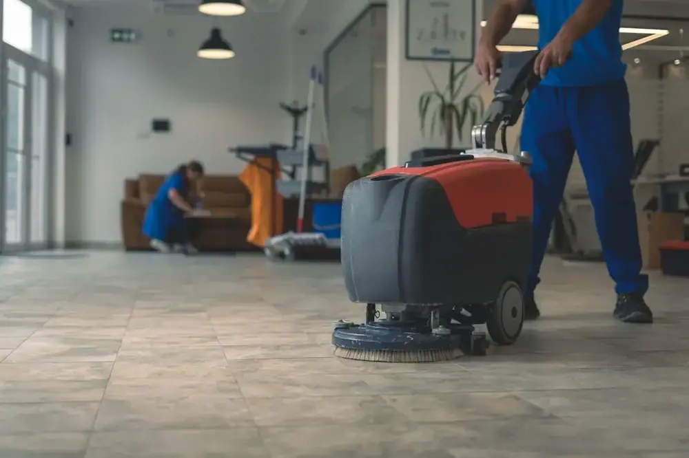 A person in blue uniform uses a floor cleaning machine on a tiled floor in a modern TX room. In the background, another person, experienced in carpet cleaning Houston County, cleans near a couch and exercise equipment.