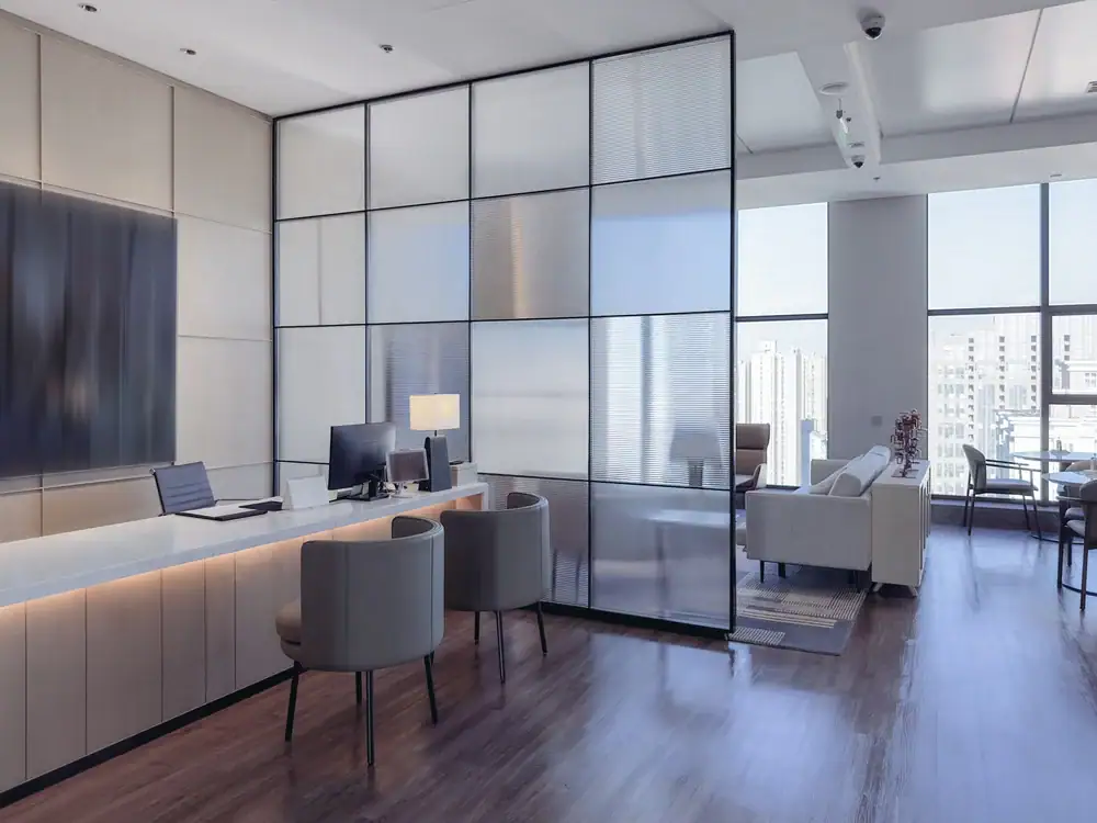 Modern office with large windows, a long reception desk, two grey chairs, a computer, and a lamp. Glass partition separates the reception area from a lounge with sofas and city views—ideal for businesses seeking carpet cleaning Houston County, TX.