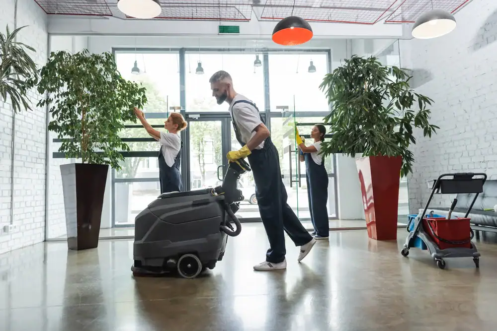 Three janitors in uniforms clean a modern, bright lobby. One uses a floor cleaning machine, while two others wipe windows. Large potted plants and cleaning supplies reflect the professional carpet cleaning Houston County, TX trusts.