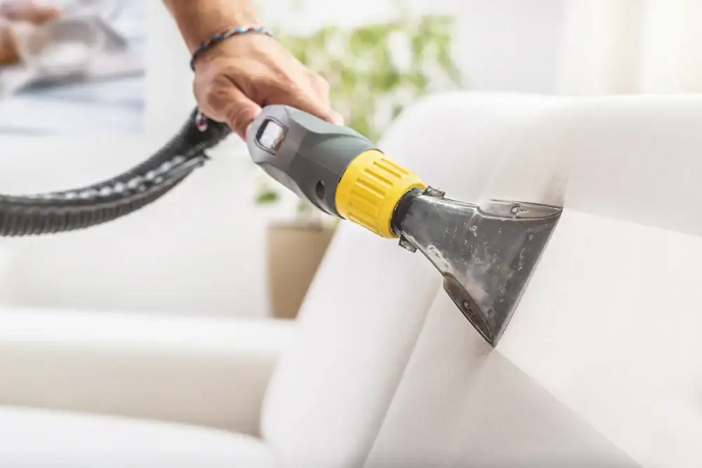 A person uses a vacuum cleaner with a narrow attachment to clean a white upholstered sofa, removing dust and dirt from the fabric—an important step in maintaining fresh furniture between carpet cleaning Houston County,TX appointments.