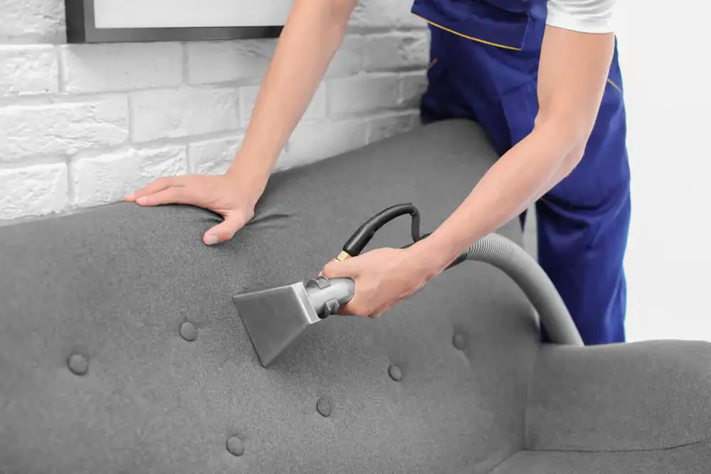 A person wearing blue overalls is using a vacuum cleaner with a special attachment to clean a gray upholstered sofa, showcasing expert carpet cleaning Houston County, TX, against a white brick wall.