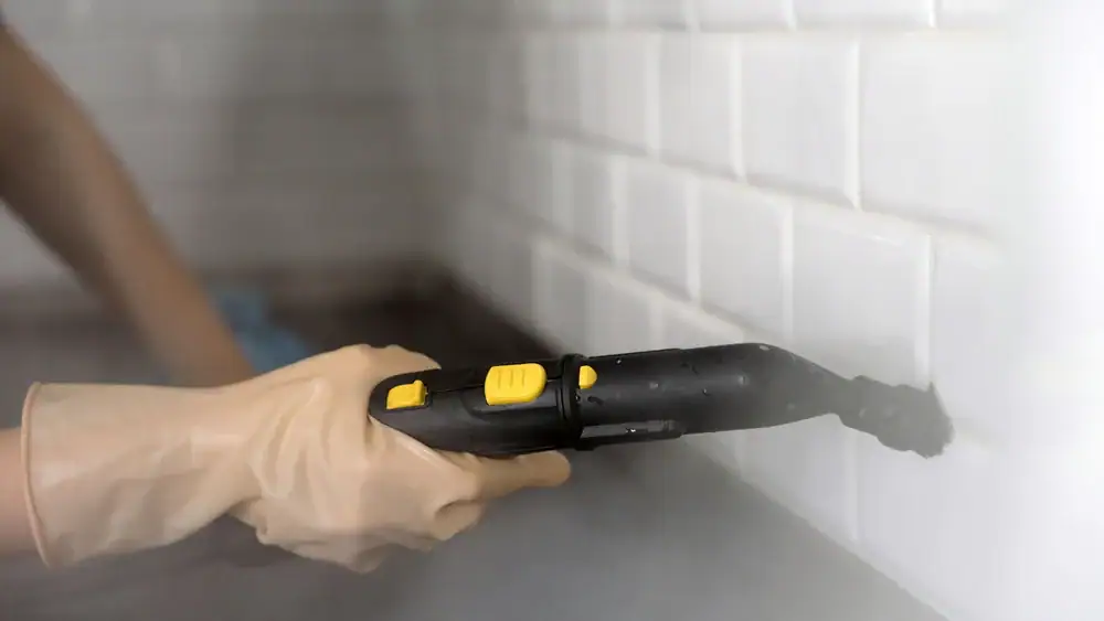 A person in TX wearing a glove uses a handheld steam cleaner to clean white tiled grout on a wall, with visible steam rising from the device—an effective technique often used by carpet cleaning Houston County experts.