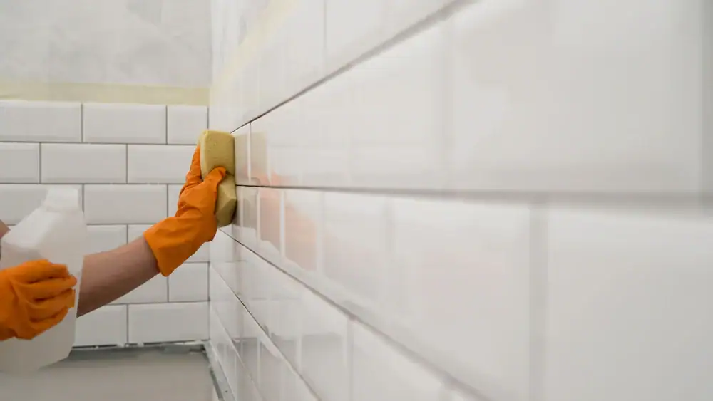 Person wearing orange gloves cleaning glossy white subway tiles in TX with a yellow sponge and holding a cleaning bottle. The evenly arranged tiles shine—perfect for those seeking more than just carpet cleaning in Houston County.