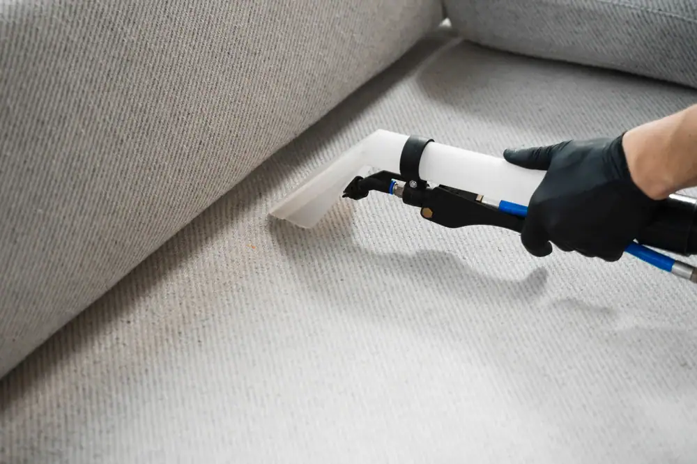 A person wearing a black glove uses a handheld vacuum or upholstery cleaning tool to clean a light gray fabric sofa, demonstrating expert carpet cleaning Houston County, TX service.