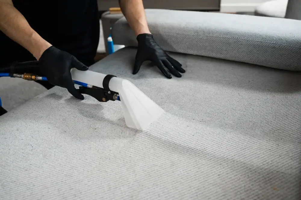 A person wearing black gloves uses a handheld upholstery cleaning machine to clean a light grey fabric sofa, demonstrating expert carpet cleaning Houston County, TX services.