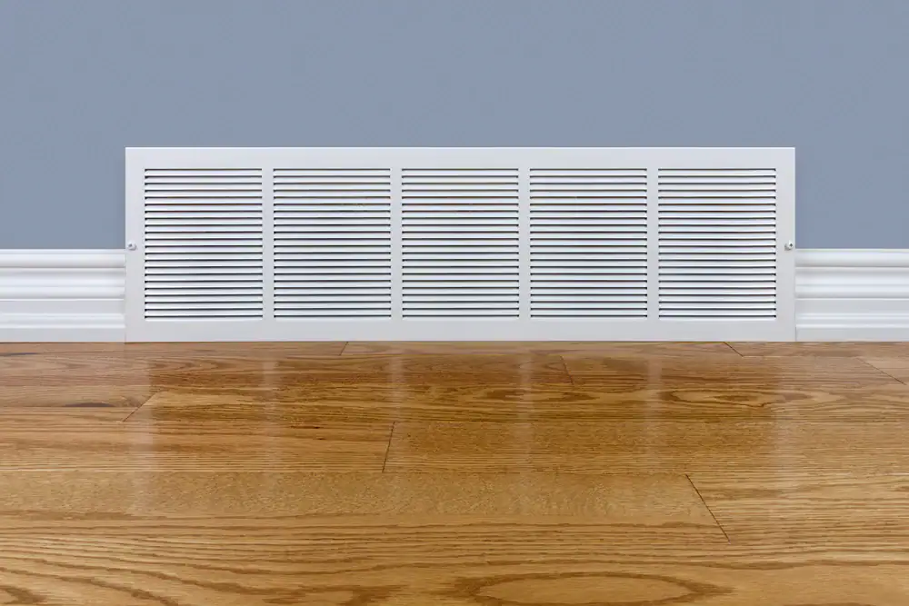 A white air vent cover is installed on a light blue wall above a wooden floor with white baseboards in TX, where quality carpet cleaning Houston County services are also available.